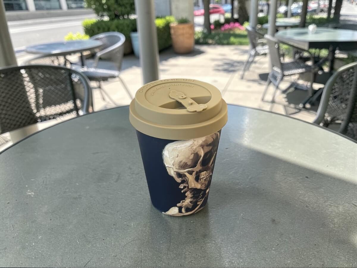 Ecoffee Cup Skull of a Skeleton Van Gogh Museum bicchiere da caffè 350ml con motivo teschio su tavolo rotondo in metallo di terrazza caffetteria con sedie metalliche e sole d'estate