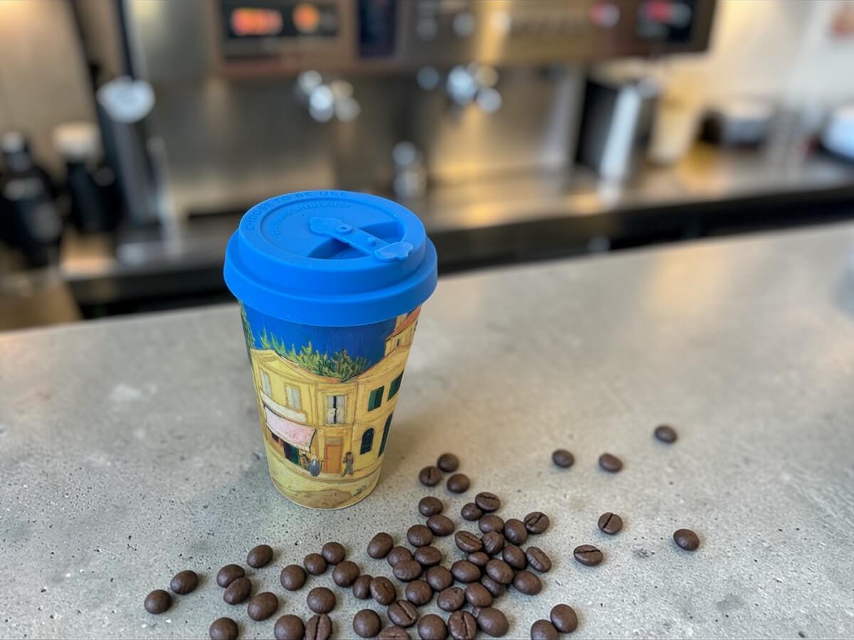 Ecoffee Cup The Yellow House 400ml Van Gogh Museum con motivo La Casa Gialla su bancone in cemento con chicchi di caffè sparsi e macchina espresso professionale sullo sfondo in caffetteria