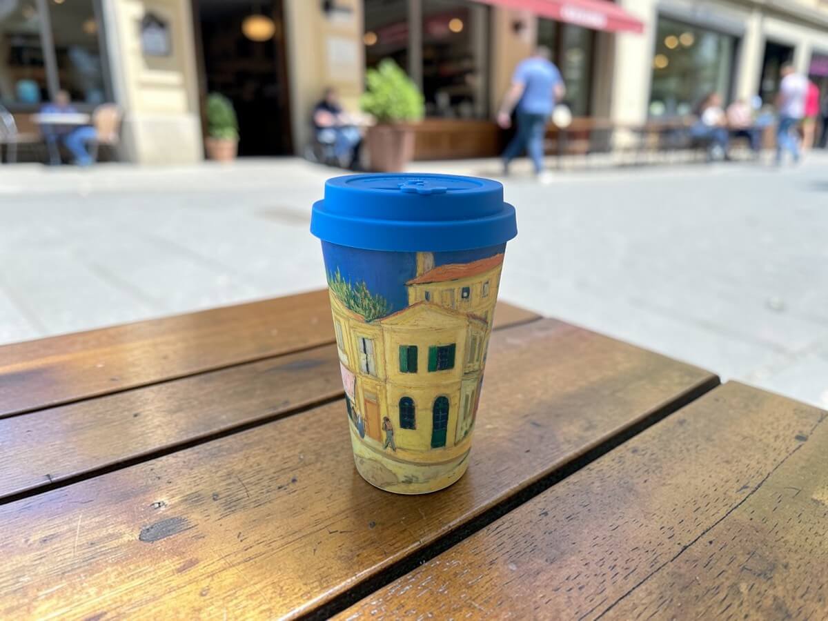 Ecoffee Cup The Yellow House 400ml Van Gogh Museum con motivo La Casa Gialla su tavolo in legno di terrazza caffetteria con scena urbana animata e passanti alla luce del sole