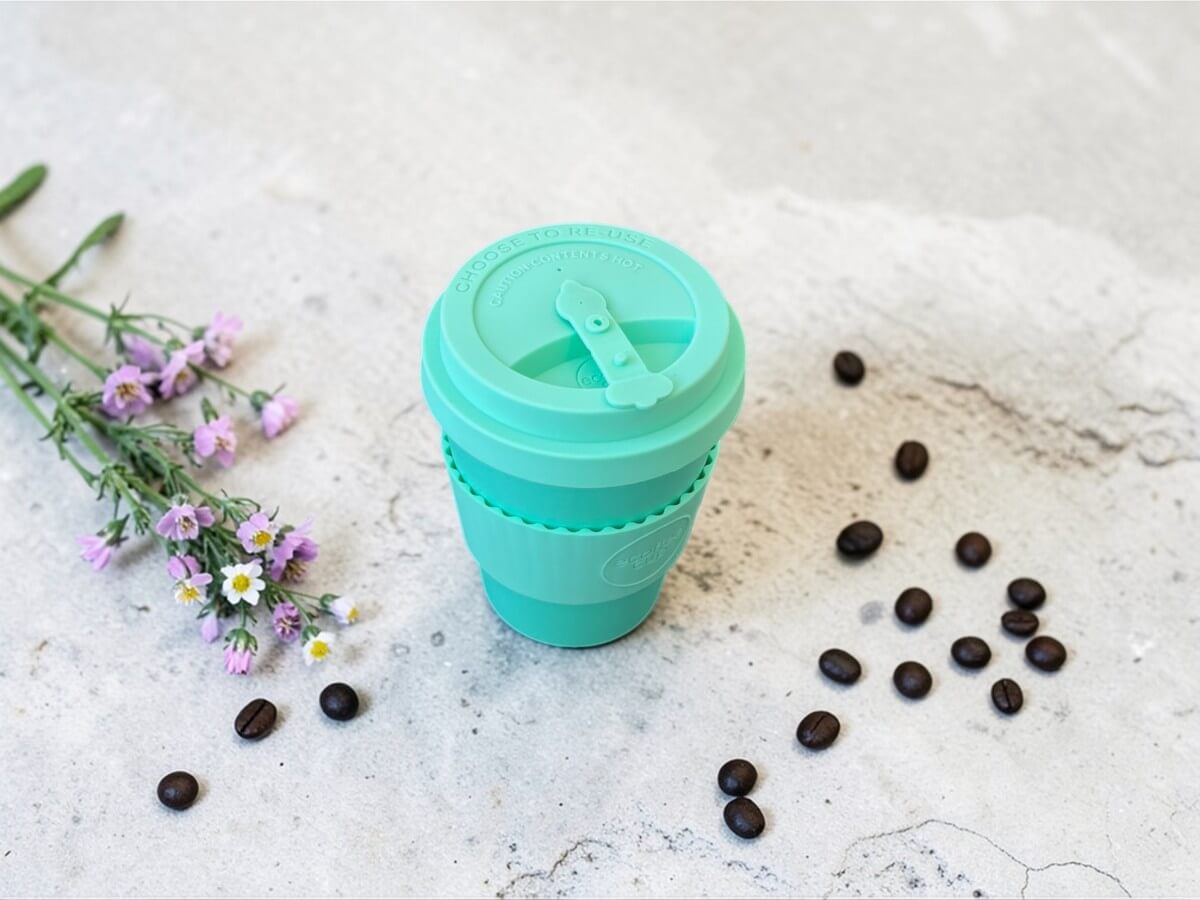 Ecoffee Cup bicchiere riutilizzabile 350ml Grindavik Split in verde menta su superficie in cemento chiaro accanto a fiori viola e chicchi di caffè sparsi alla luce naturale
