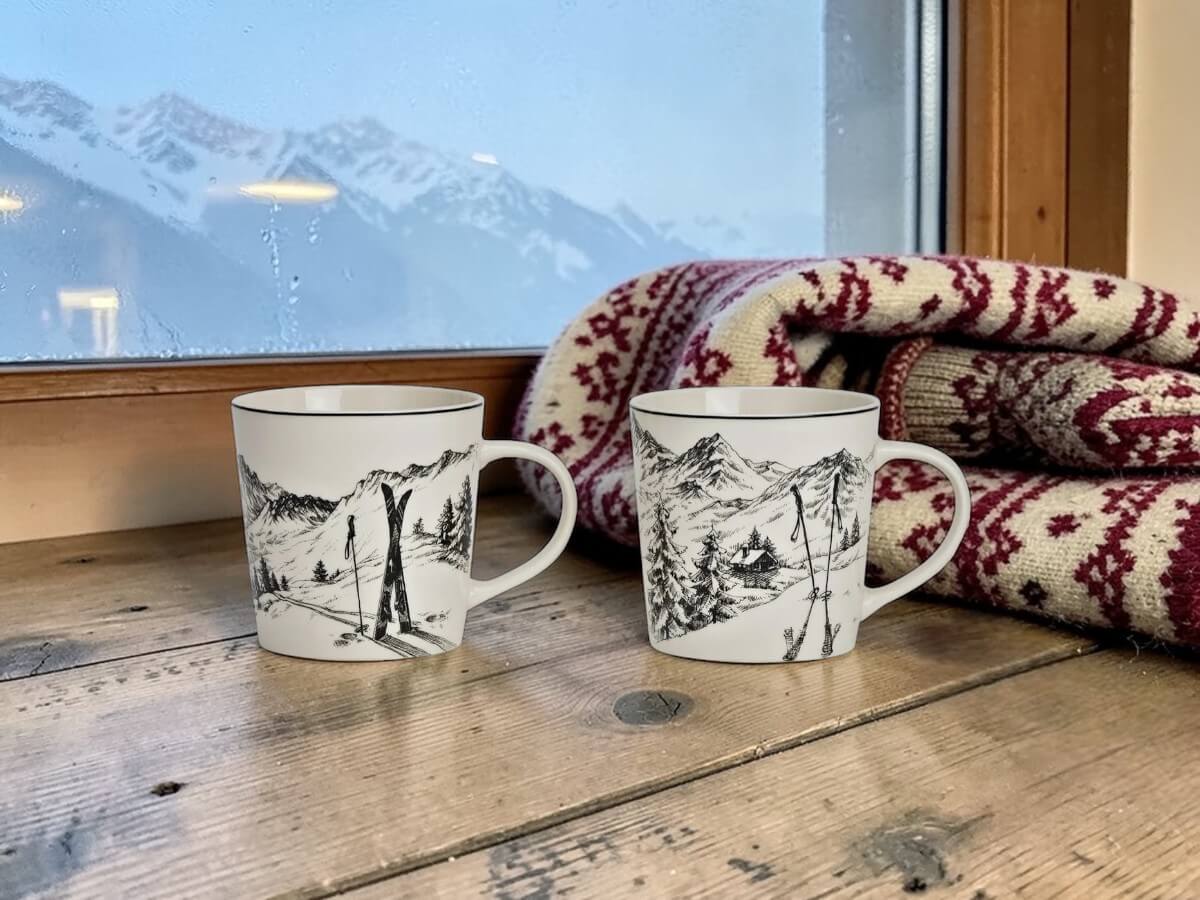 Zwei Porzellanbecher Gipfelrast mit Ski- und Alpendekor auf rustikaler Holzfensterbank in Alpenchalet mit traditioneller Schweizer Strickdecke in Rot und Weiss und verschneiten Berggipfeln durch beschlagenes Fenster