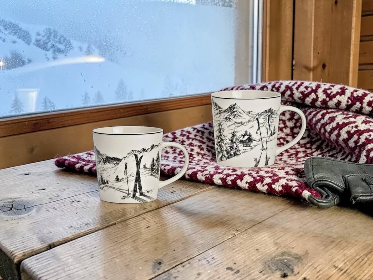 Zwei Porzellanbecher Gipfelrast mit Ski- und Alpendekor auf rustikaler Holzfensterbank in Schweizer Berghütte mit rot-weisser Wolldecke Lederhandschuhen und verschneitem Bergpanorama durch beschlagenes Fenster