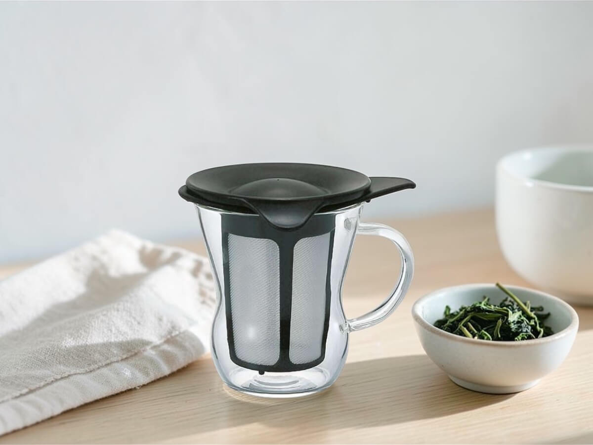 Hario Teebereiter aus Glas fuer eine Tasse mit integriertem Filter auf hellem Holztisch mit losem Tee im Lifestyle-Setting.