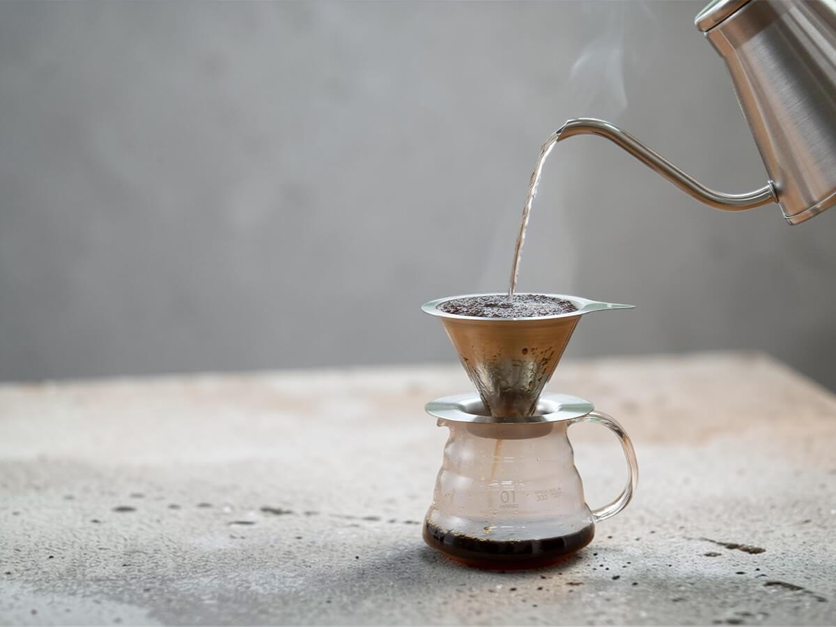 Hario V60 Double Mesh Metallfilter 01 aus Edelstahl beim Handaufguss mit Gooseneck Wasserkocher auf Glaskanne auf heller Steinoberflaeche