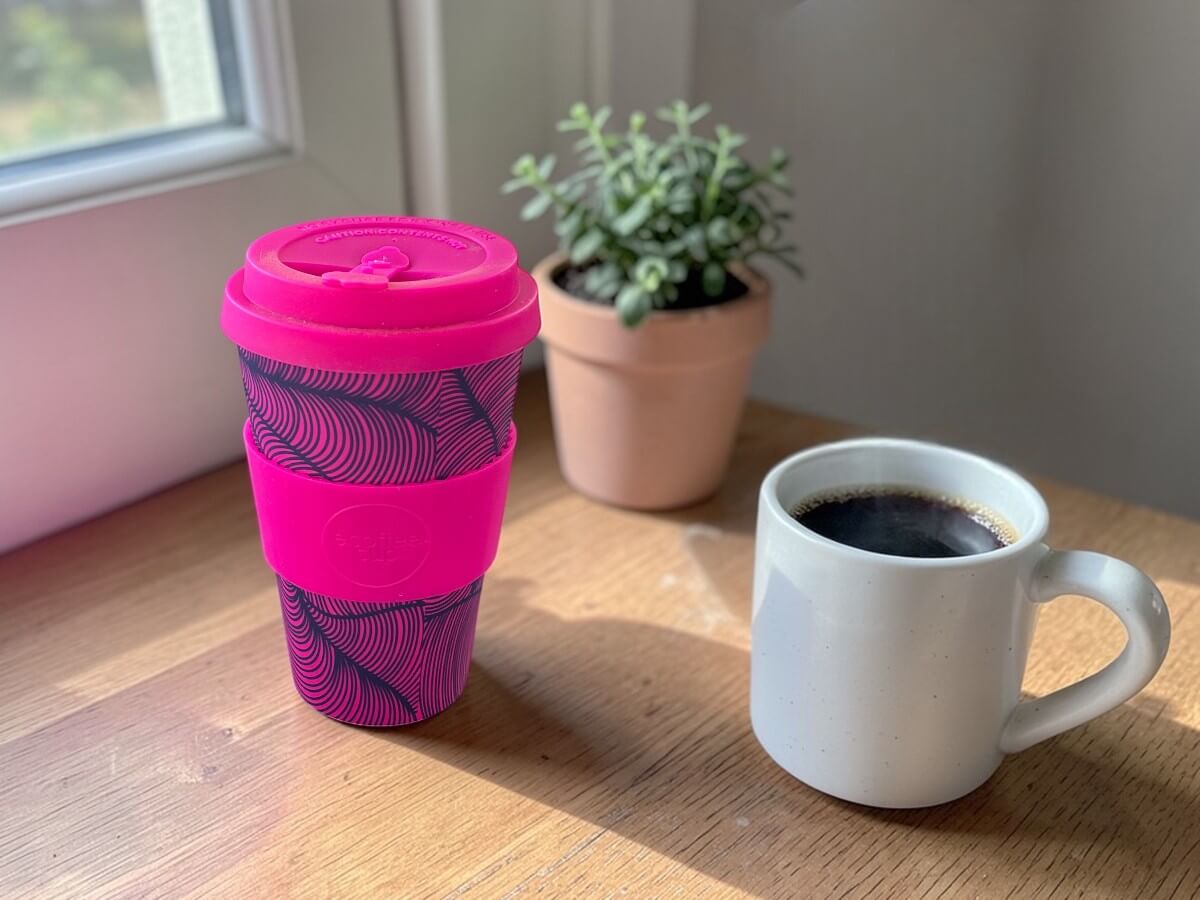 Tazza da caffè in PLA 400ml Otrobanda in magenta su tavolo in legno accanto a tazza da caffè in ceramica con caffè nero e succulenta in vaso di terracotta vicino alla finestra nella calda luce del mattino