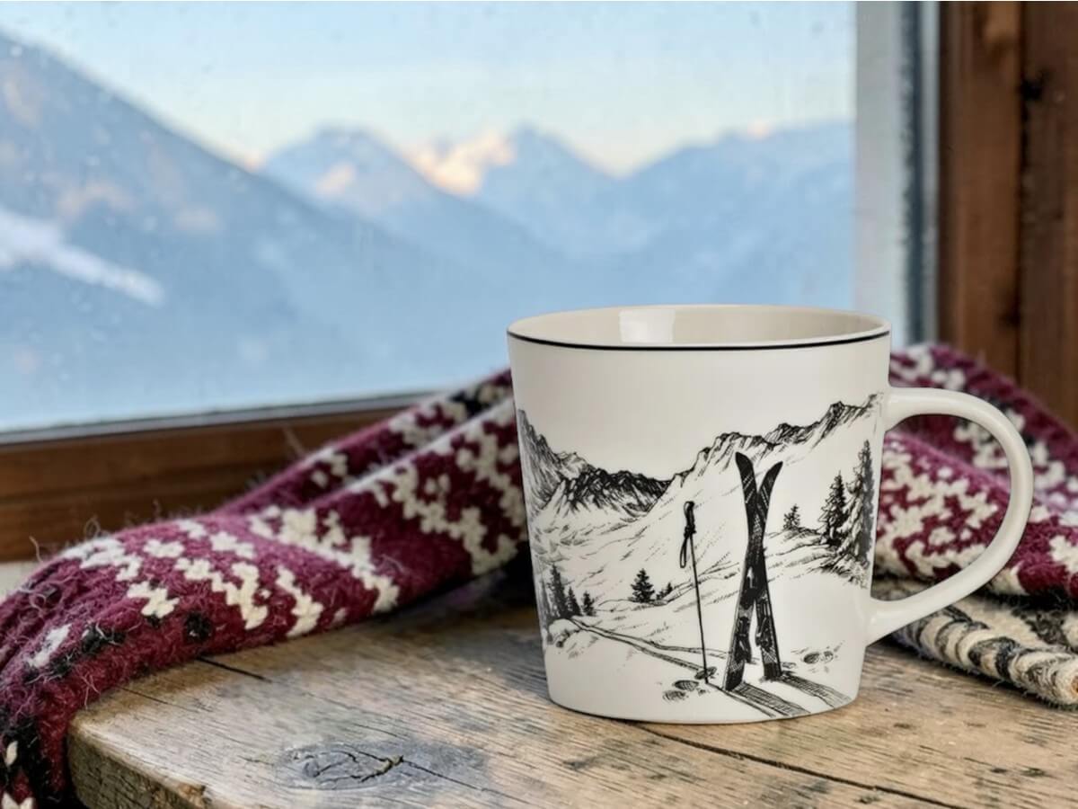 Porcelain mug Pulverschnee with ski and mountain panorama pattern on rustic wooden windowsill in mountain lodge with red and white Norwegian pattern wool blanket and alpine mountain range in evening light through frosted window