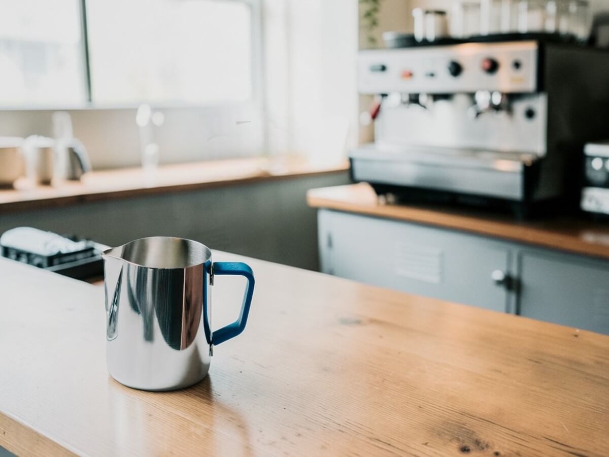 Rhino Blue Silikon Griffüberzug 12oz an Milchkännchen auf Holztisch mit Espressomaschine im Hintergrund