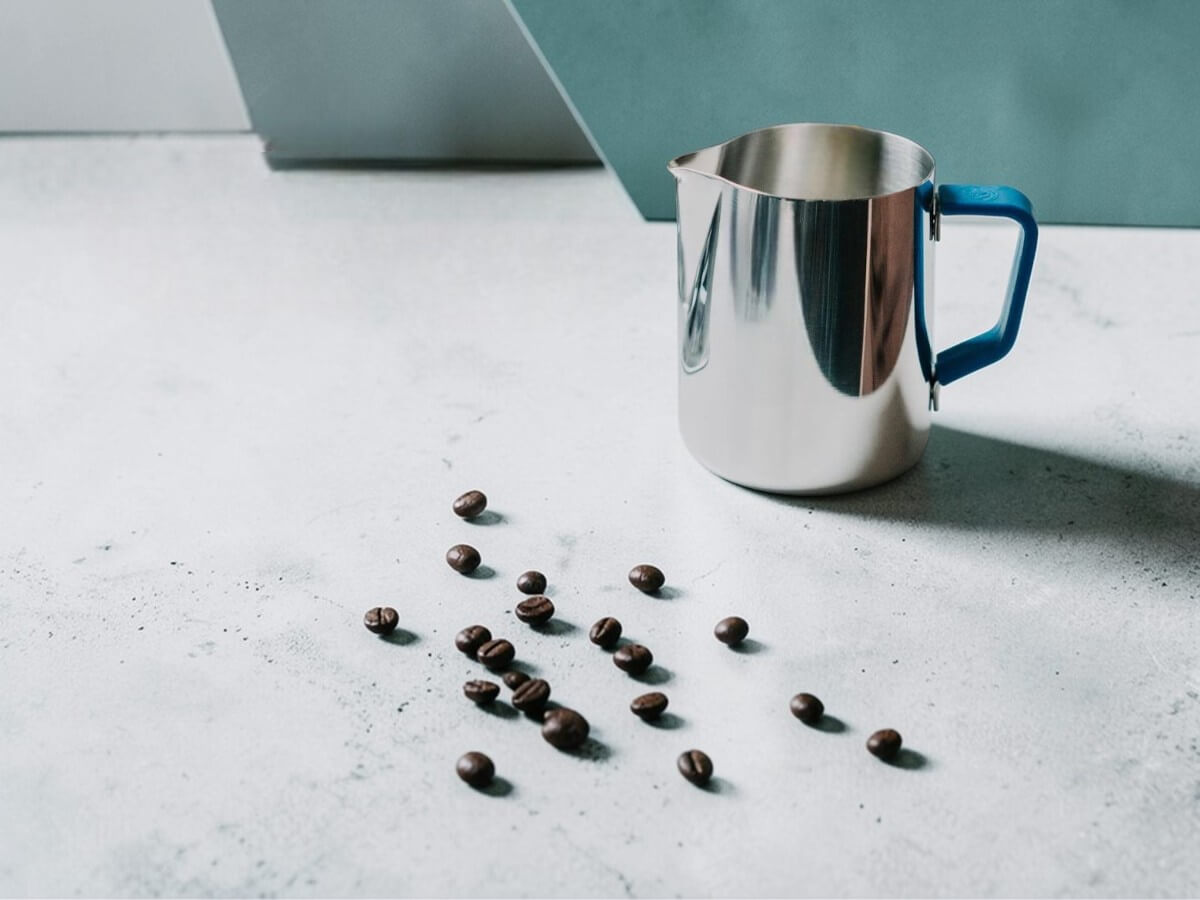 Rhino Blue Silikon Griffüberzug 32oz an Milchkännchen auf Betonoberfläche mit verstreuten Kaffeebohnen