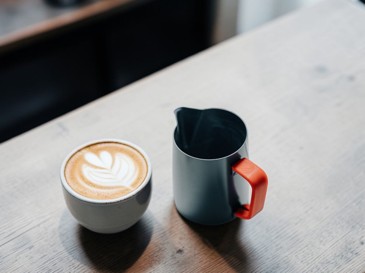 Rhino Red Silikon Griffüberzug 32oz an Milchkännchen neben Cappuccino mit Latte Art auf Holztisch