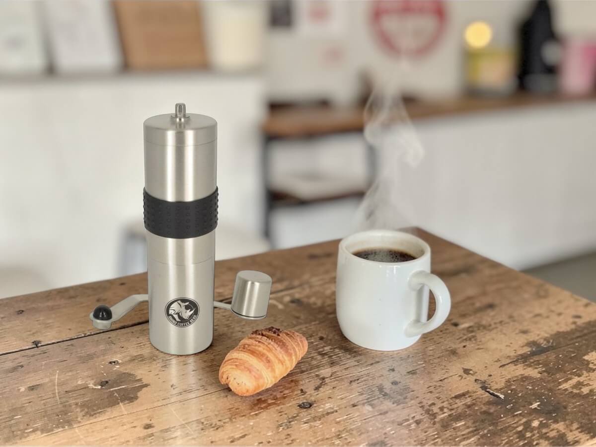 Moulin à café manuel Rhino en acier inoxydable sur table en bois à côté d'une tasse de café chaud et croissant
