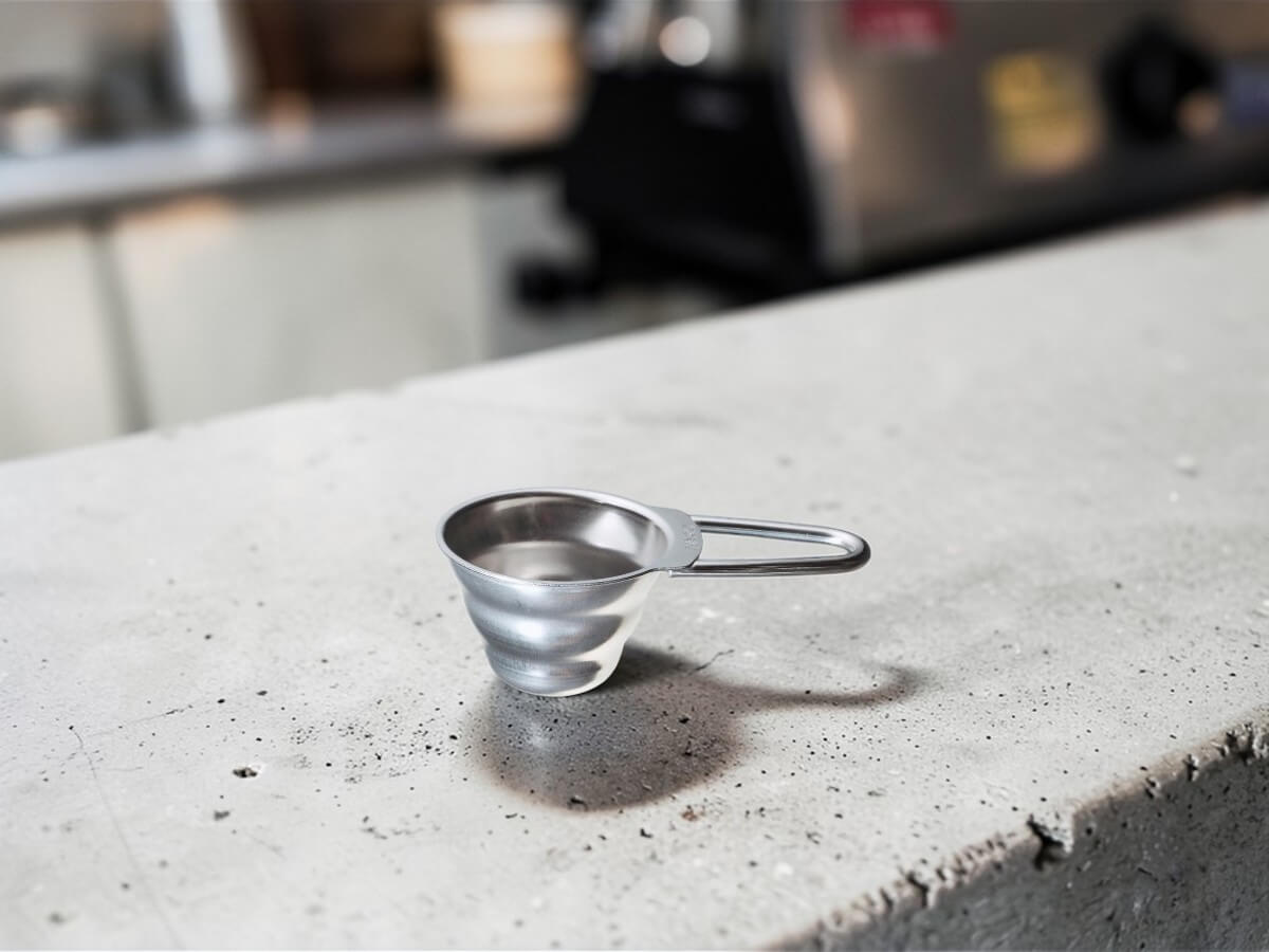 V60 Messlöffel 12g aus Edelstahl auf Betonoberfläche im Café mit Kaffeemaschine im Hintergrund