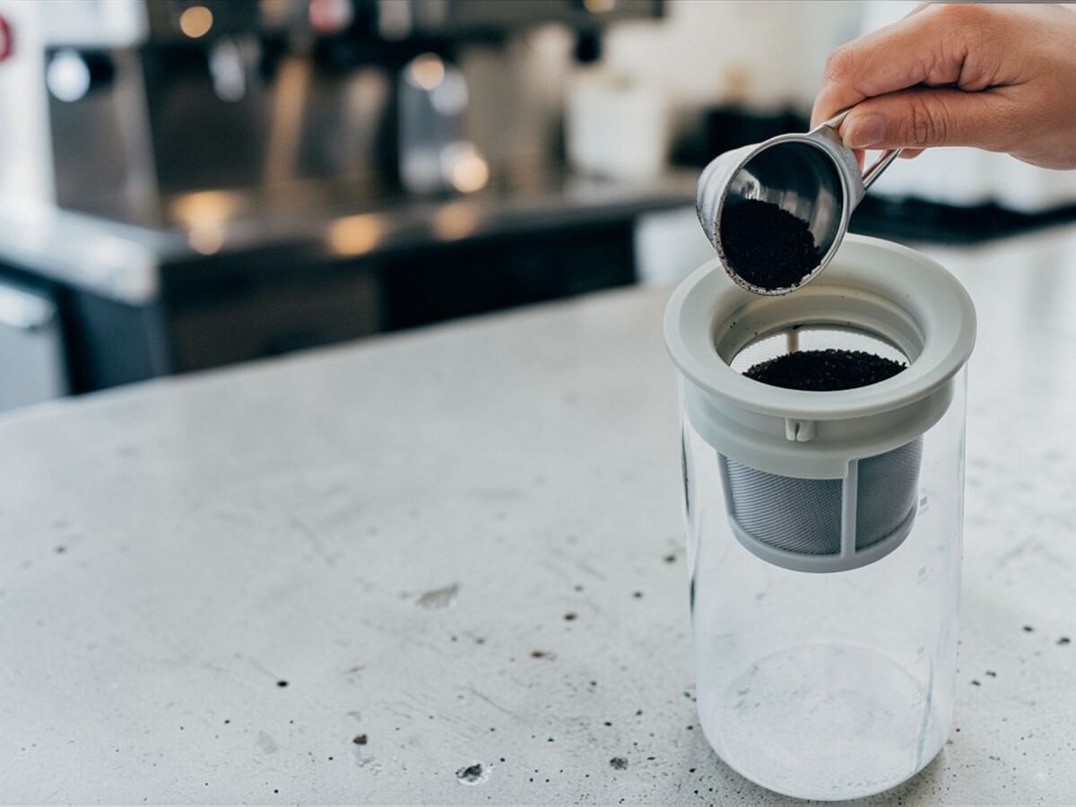 V60 Messlöffel 12g aus Edelstahl beim Dosieren von Kaffeepulver in Glasbehälter mit Filtereinsatz