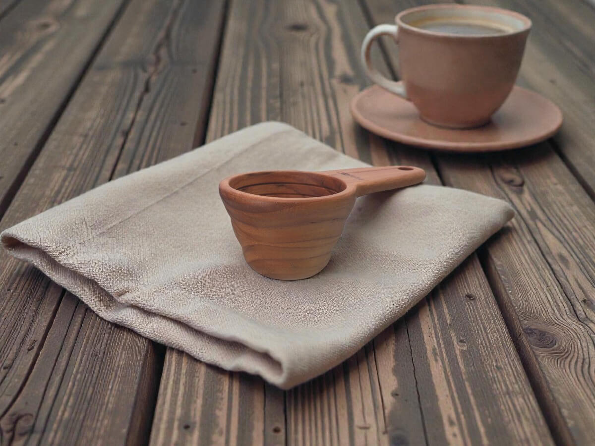 V60 Messlöffel 12g aus Holz auf Leinentuch auf rustikalem Holztisch mit Kaffeetasse im Hintergrund